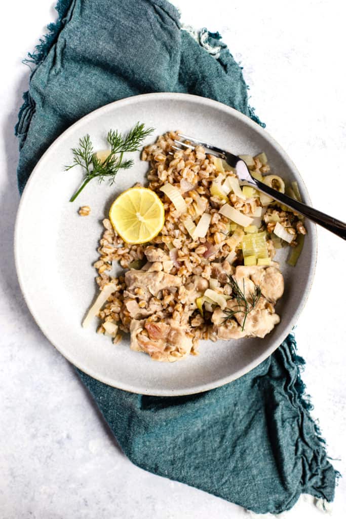 One skillet chicken with leeks and farro in light blue serving bowl with slice of lemon and fresh dill for serving and fork resting in bowl, on teal linen on blue and white surface