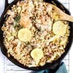 One skillet chicken with leeks and farro in cast iron skillet fresh out of the oven with lemon slices on top, wooden spoon, teal linen around the handle, on cooling rack, on white and blue surface