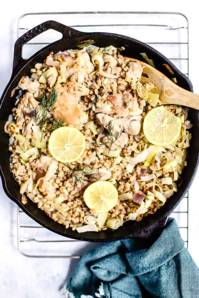 One skillet chicken with leeks and farro in cast iron skillet fresh out of the oven with lemon slices on top, wooden spoon, teal linen around the handle, on cooling rack, on white and blue surface