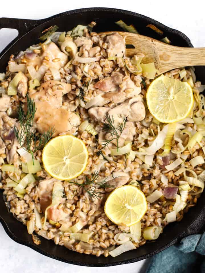 One skillet chicken with leeks and farro in cast iron skillet fresh out of the oven with lemon slices on top, wooden spoon, teal linen around the handle, on white and blue surface