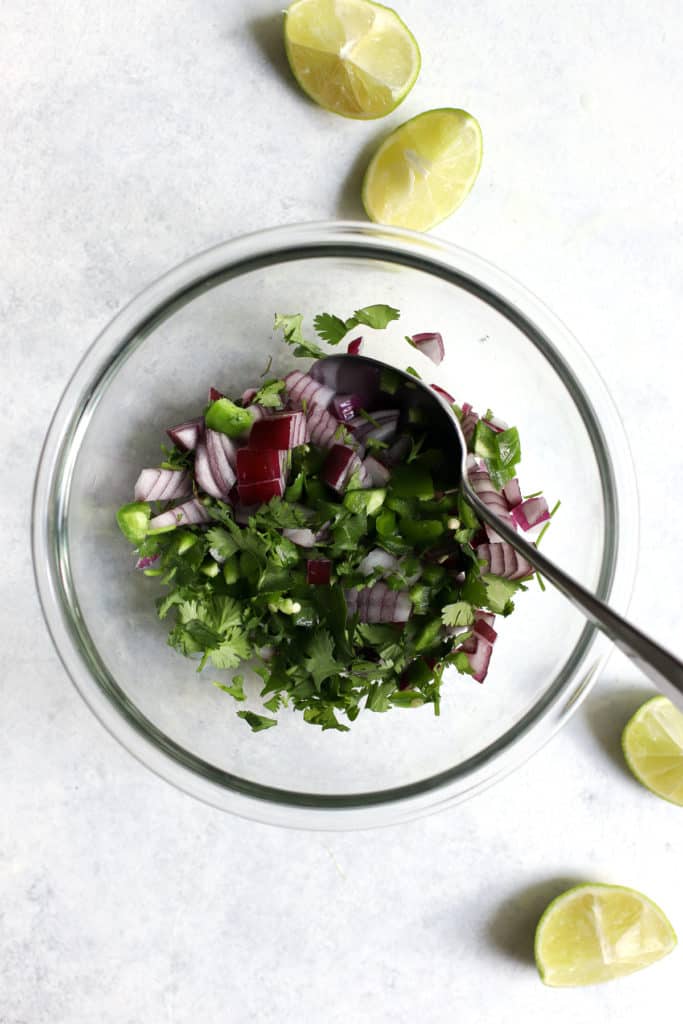 Diced red onion, jalapeños, and cilantro in glass bowl with spoon.