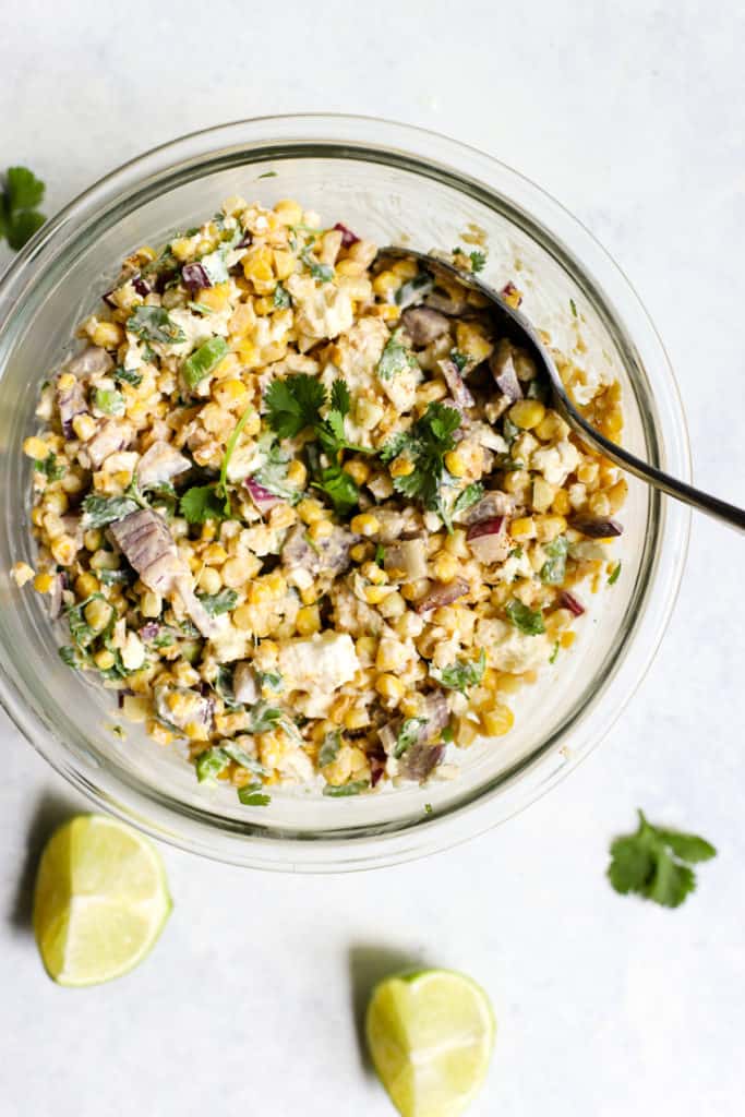 20-minute healthy street corn in a clear glass bowl with silver spoon on blue and white surface with two lime wedges