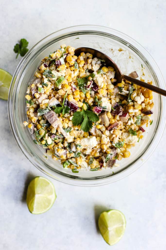 20-minute healthy street corn in a clear glass bowl with silver spoon on blue and white surface with two lime wedges