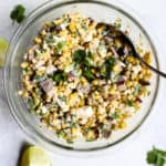 20-minute healthy street corn in a clear glass bowl with silver spoon on blue and white surface with two lime wedges