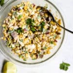 20-minute healthy street corn in a clear glass bowl with silver spoon on blue and white surface with two lime wedges