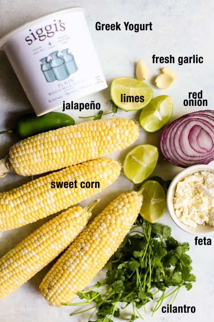 20-minute healthy street corn ingredients on blue and white surface: sweet corn on the cob, jalapeño, cilantro, lime, garlic, Greek yogurt, red onion, lime wedges, and feta