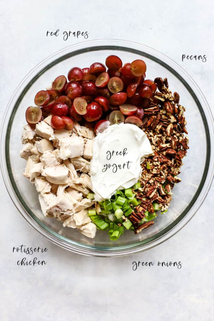 Rotisserie chicken pieces, halved red grapes, Greek yogurt, sliced green onions, and toasted pecans in clear glass bowl.