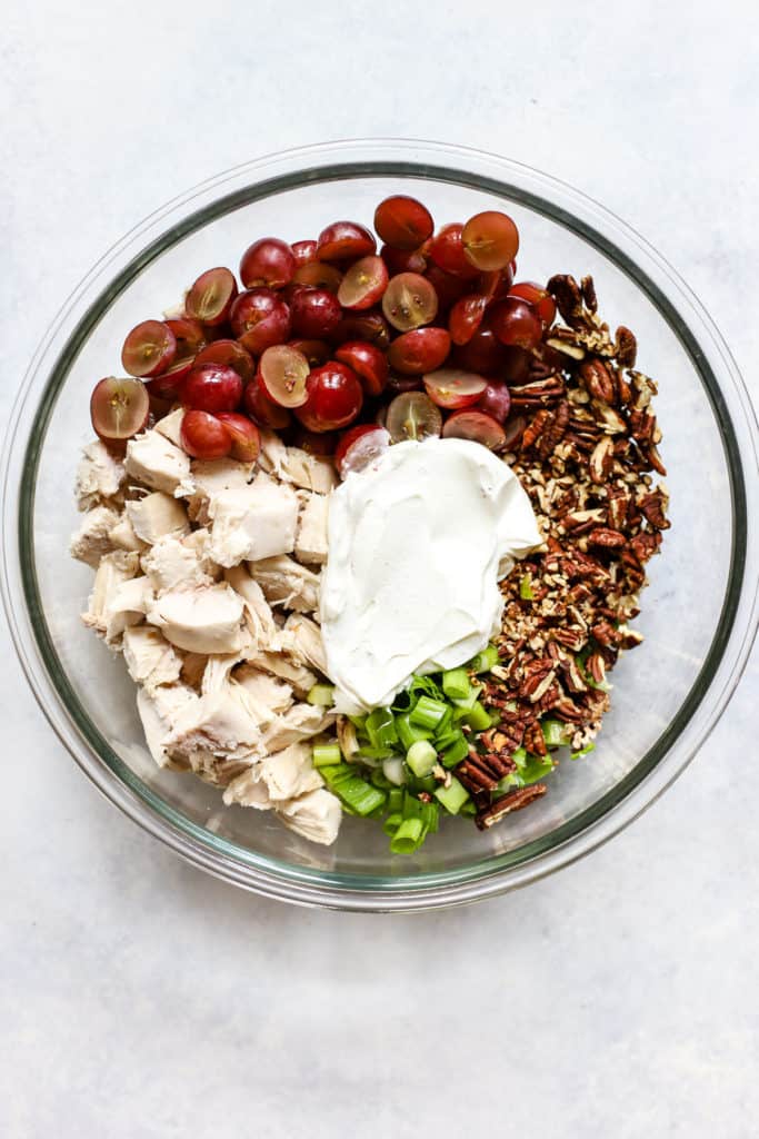 Rotisserie chicken pieces, halved red grapes, Greek yogurt, sliced green onions, and toasted pecans in clear glass bowl, before being combined.