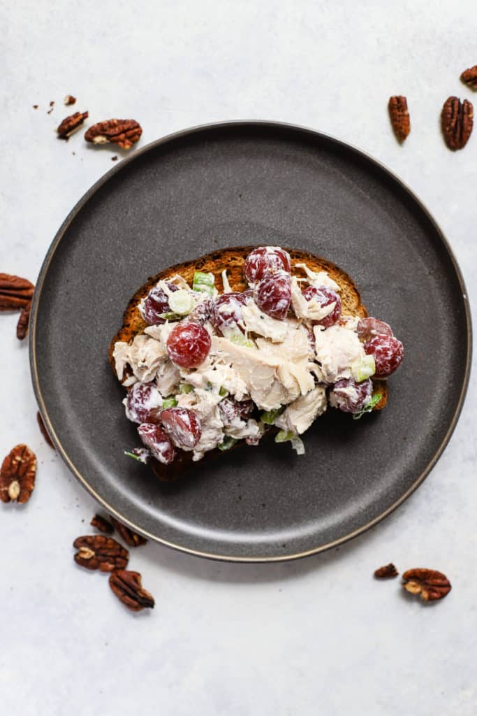 Chicken salad with grapes and pecans on toasted whole grain bread, on dark gray plate with pecans sprinkled around.