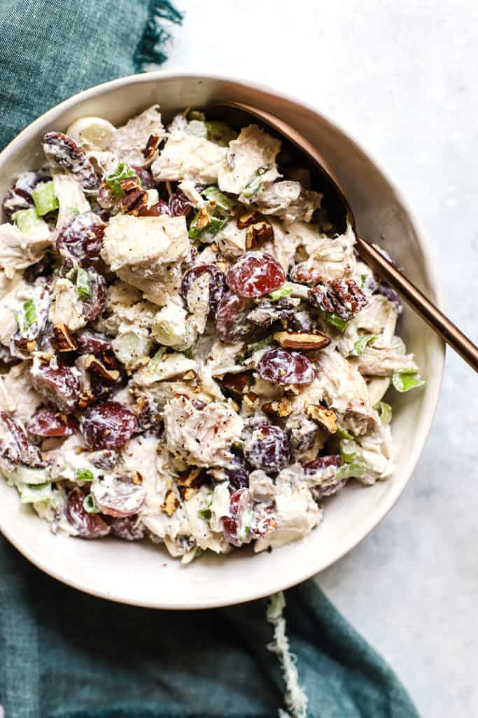 Chicken salad with grapes and pecans in white ceramic bowl with copper serving spoon on teal linen, on white and light blue surface