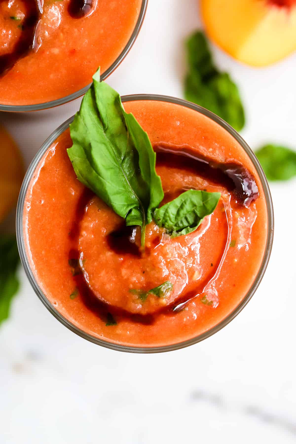 Two glasses of peach basil gazpacho with balsamic glaze swirl on top and basil leaf garnish, on white marble surface