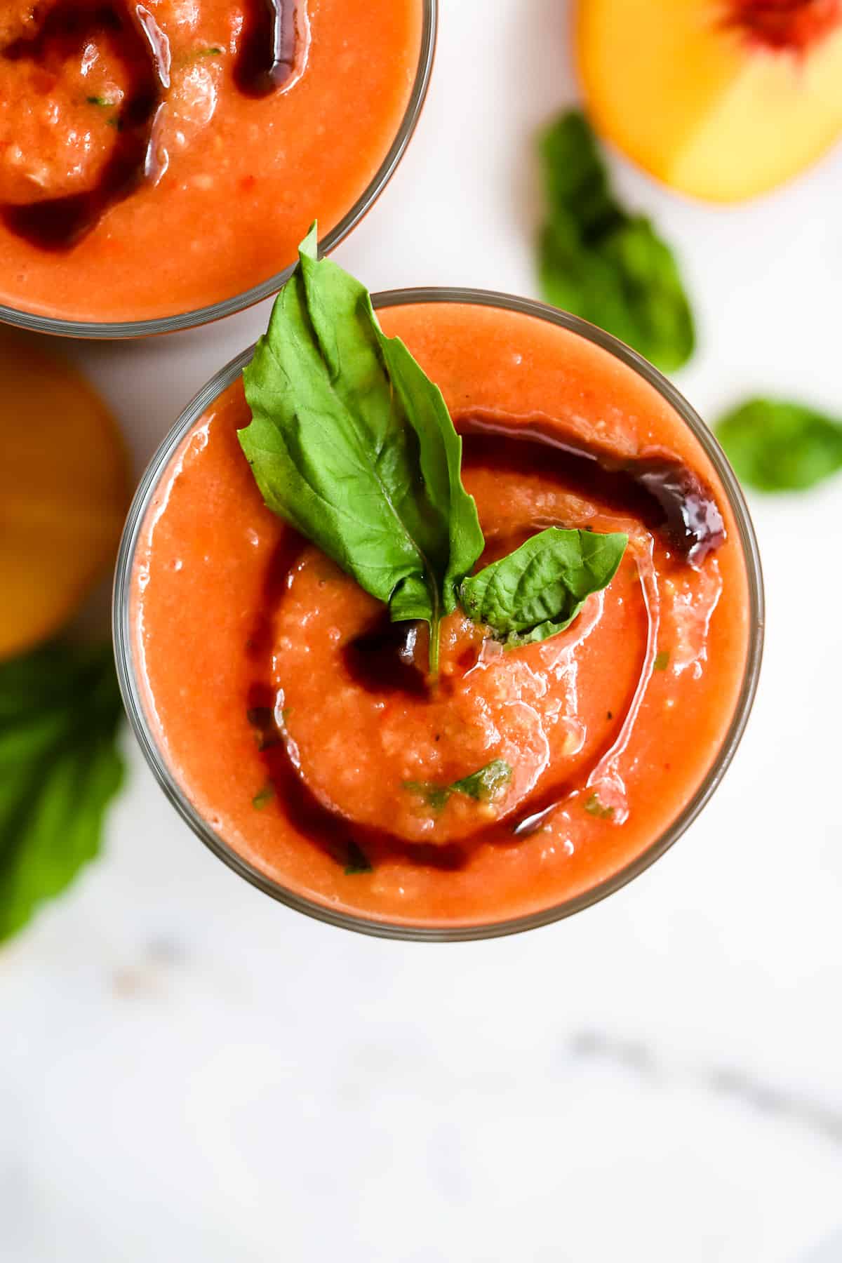 Two glasses of peach basil gazpacho with balsamic glaze swirl on top and basil leaf garnish, on white marble surface