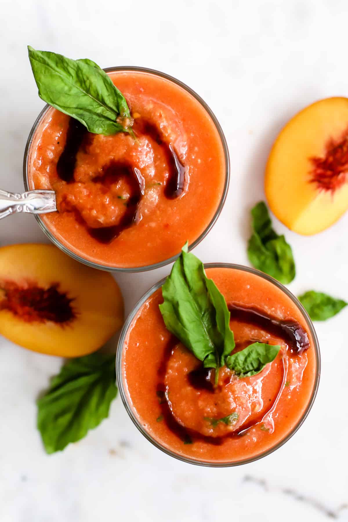 Two glasses of peach basil gazpacho with balsamic glaze swirl on top and basil leaf garnish, peach wedges on the side, on white marble surface