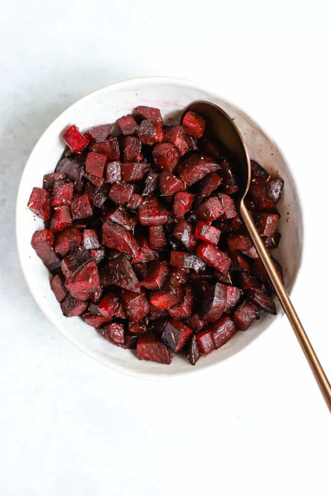 Simple herb roasted beets in white ceramic bowl with copper serving spoon on light blue background