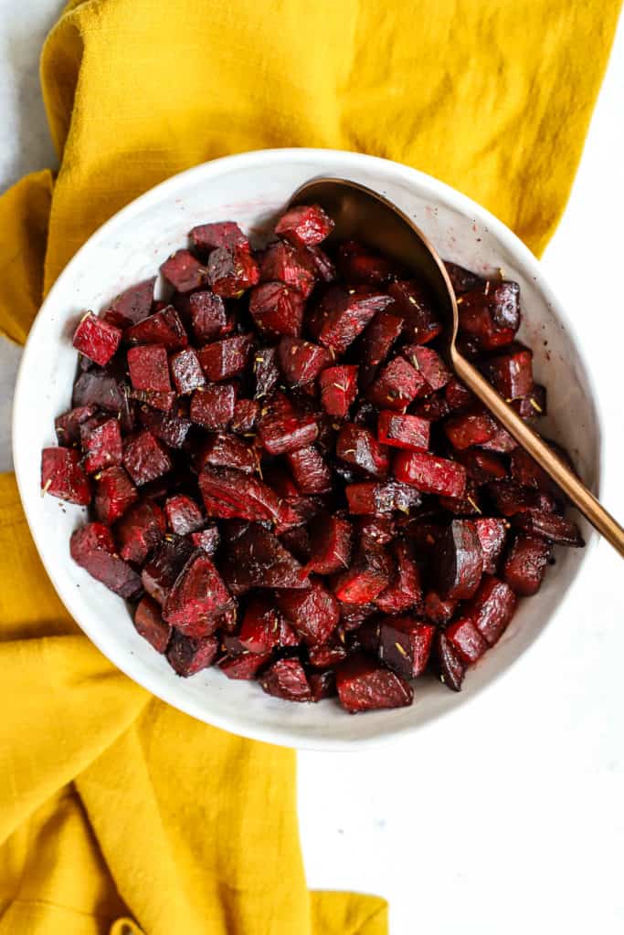 Simple herb roasted beets in white ceramic bowl with copper serving spoon on light blue background with golden yellow linen