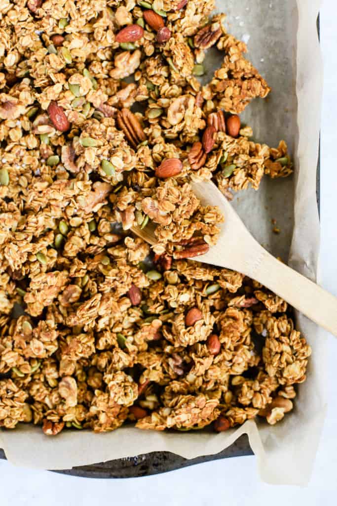 Healthy pumpkin granola in parchment paper-lined sheet pan with wooden spoon stirring the granola cluster