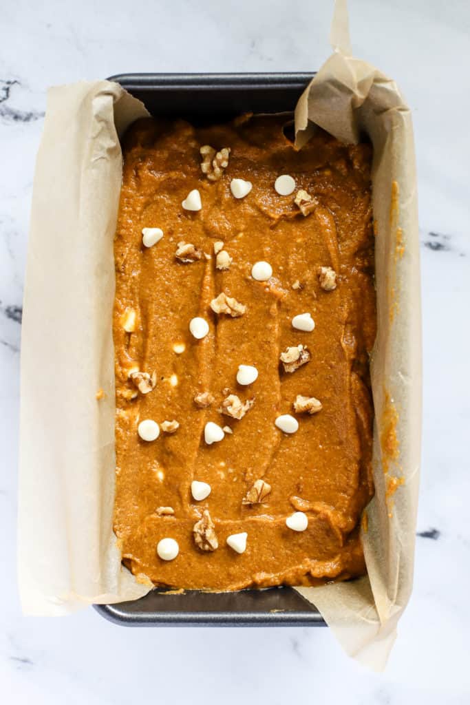 Healthy spelt pumpkin bread batter spread evenly into bread pan lined with parchment paper, with extra white chocolate chips and walnuts sprinkled on top