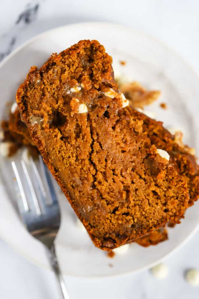 Stack of healthy spelt pumpkin bread slices on a small white plate with fork, on white marble surface, with a few walnuts and white chocolate chips sprinkled on the side