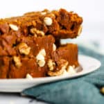 Stack of healthy spelt pumpkin bread slices on a small white plate with fork, on teal linen on white marble surface, with a few walnuts and white chocolate chips sprinkled on the side