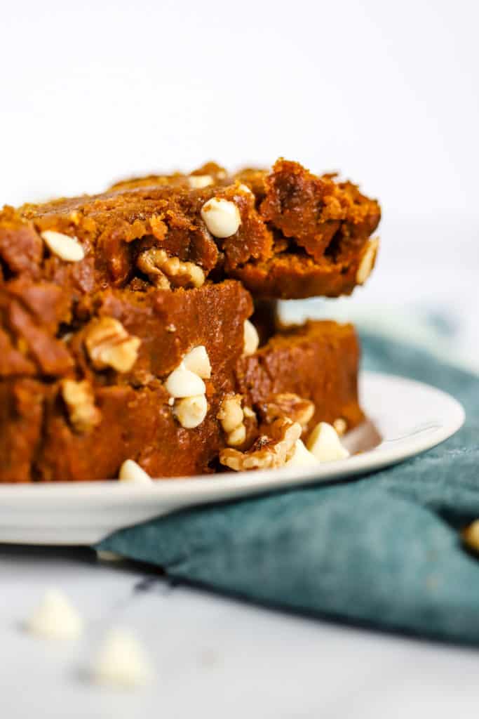 Stack of healthy spelt pumpkin bread slices on a small white plate with fork, on teal linen on white marble surface, with a few walnuts and white chocolate chips sprinkled on the side