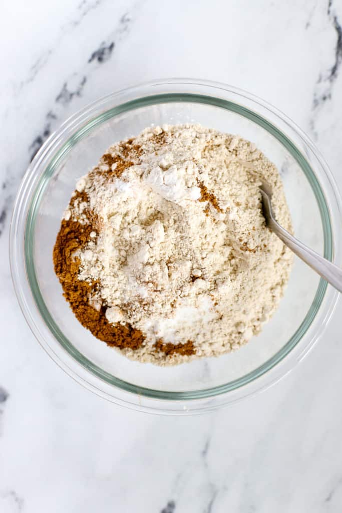 Flour, salt, baking soda, and pumpkin pie spice in small clear glass bowl with fork for whisking, on white marble surface