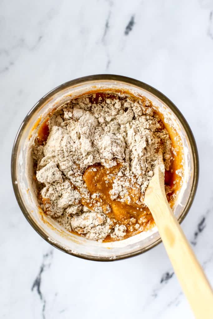 Healthy spelt pumpkin bread batter being stirred together in clear glass mixing bowl with wooden spoon, on white marble surface