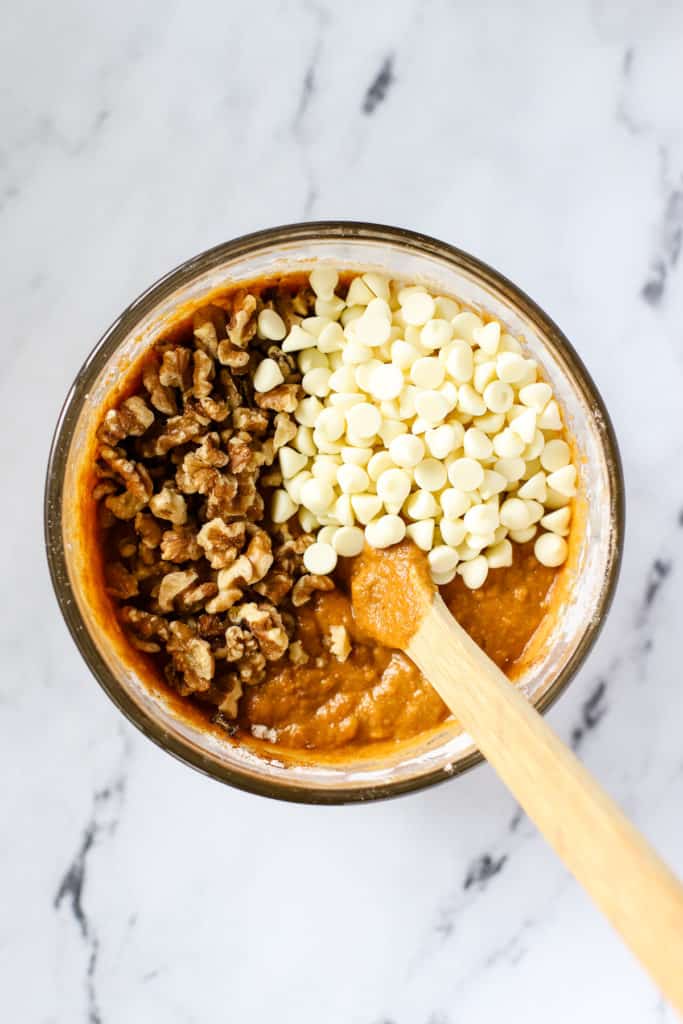 Healthy spelt pumpkin bread batter in clear glass mixing bowl with walnuts and white chocolate chips poured on top before mixing in, with wooden spoon resting in batter