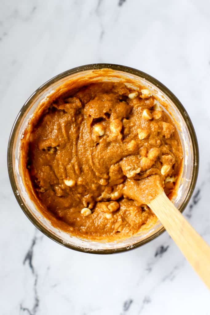 Healthy spelt pumpkin bread batter in clear glass mixing bowl with walnuts and white chocolate chips stirred in with wooden spoon, on white marble surface