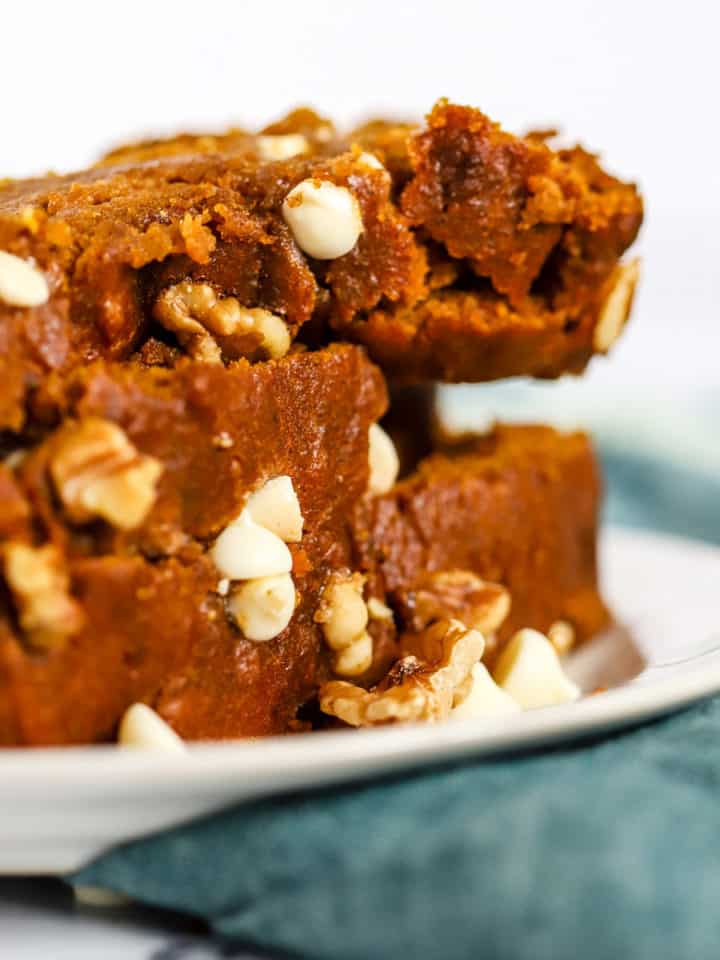 Stack of healthy spelt pumpkin bread slices on a small white plate with fork, on teal linen on white marble surface, with a few walnuts and white chocolate chips sprinkled on the side