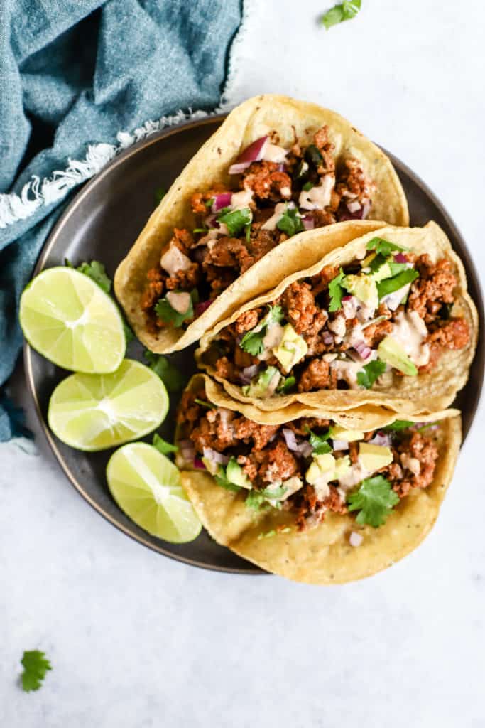 Three chorizo tacos and three lime wedges on gray plate, on light blue and white surface with teal linen