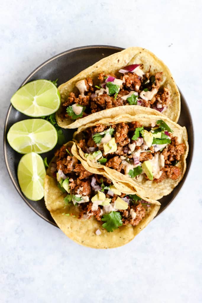 Three chorizo tacos and three lime wedges on gray plate, on light blue and white surface