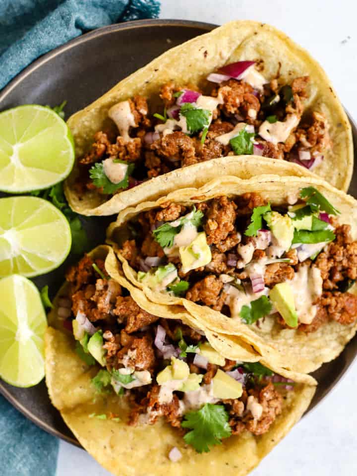 Three chorizo tacos and three lime wedges on gray plate, on light blue and white surface