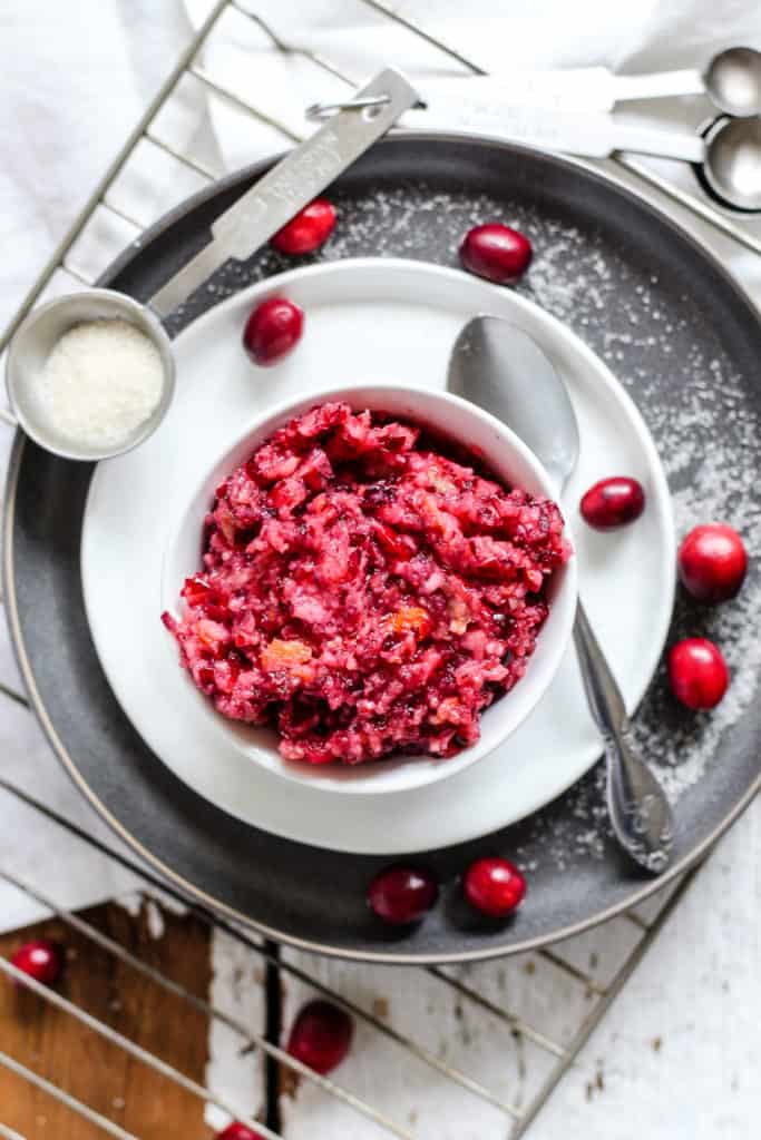 Cranberry relish in small white dish on small white plate on larger gray plate, with silver spoon and a few whole cranberries on the side