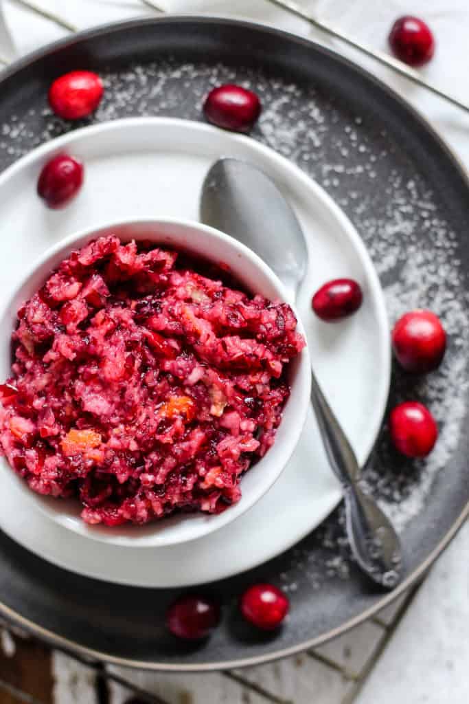 Cranberry relish in small white dish on small white plate on larger gray plate, with silver spoon and a few whole cranberries on the side