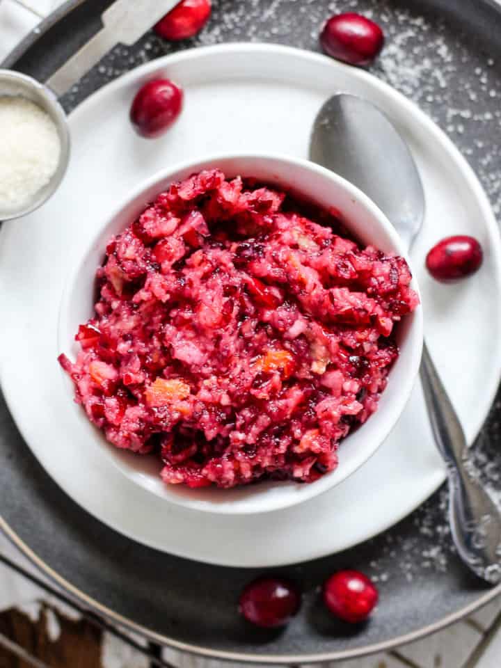 Cranberry relish in small white dish on small white plate on larger gray plate, with silver spoon and a few whole cranberries on the side