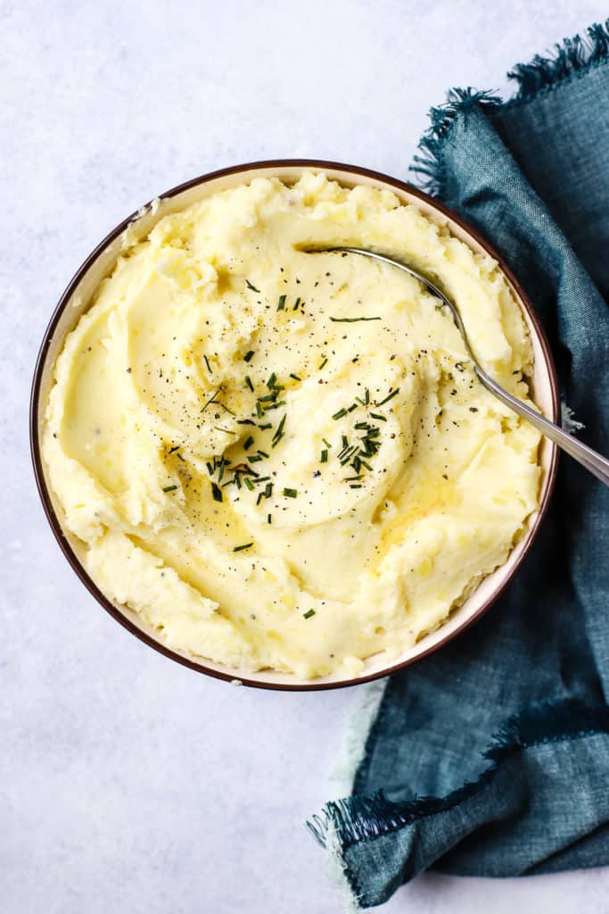 Healthy Instant Pot mashed potatoes in beige bowl with butter melted on top, chives, and pepper, with spoon & teal linen on side.