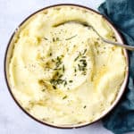 Healthy Instant Pot mashed potatoes in beige bowl with butter melted on top, chives, and pepper, with spoon & teal linen on side.