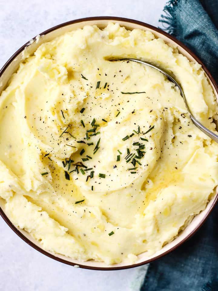 Healthy Instant Pot mashed potatoes in beige bowl with butter melted on top, chives, and pepper, with spoon & teal linen on side.