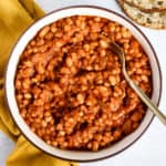 Homemade maple baked beans in beige serving bowl with gold spoon, next to golden-yellow linen, and sourdough on the side