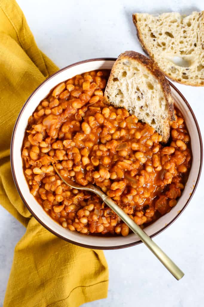 Homemade maple baked beans in beige serving bowl with gold spoon, next to golden-yellow linen, and sourdough dipped in the beans.