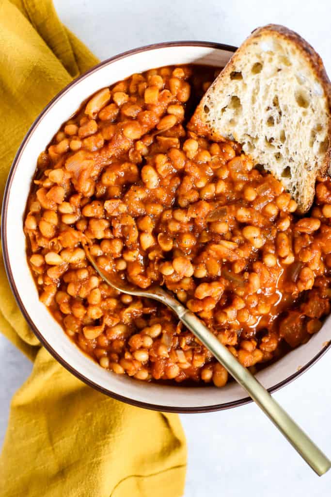 Homemade maple baked beans in beige serving bowl with gold spoon, next to golden-yellow linen, and sourdough dipped into the beans.