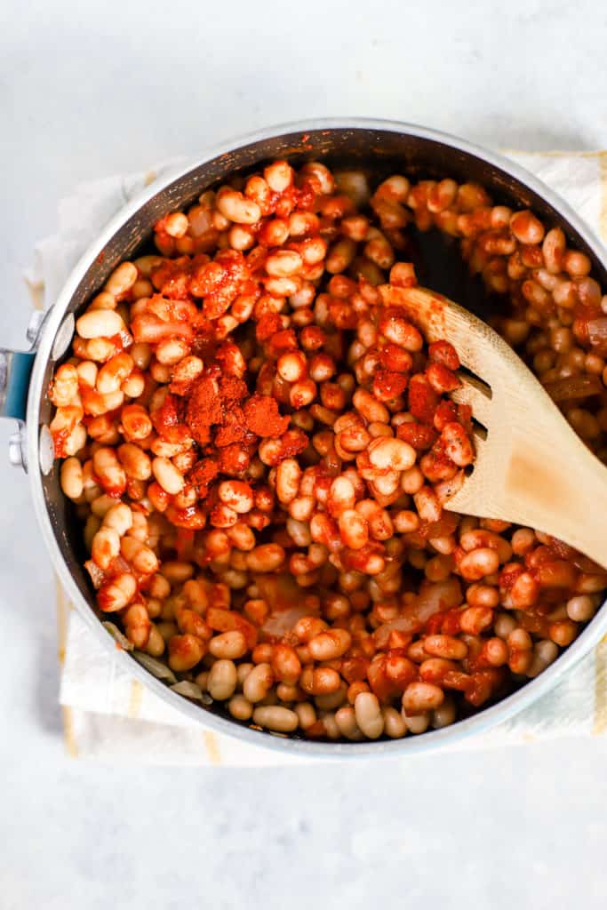 Navy beans, smoked paprika, tomato paste, onions, and garlic in small pot with bamboo stirring spoon.