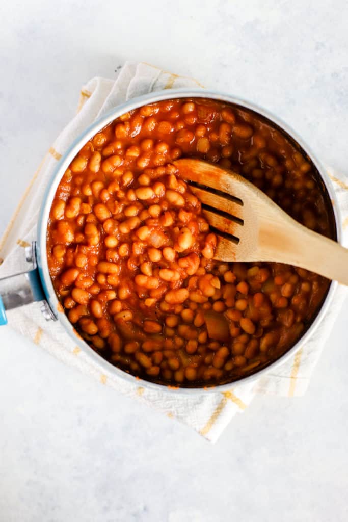 Homemade maple baked beans in small blue pot, done cooking, with bamboo spoon. The pot is resting on a yellow and white windowpane print towel.