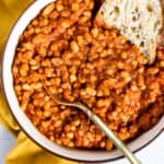 Homemade maple baked beans in beige serving bowl with gold spoon, next to golden-yellow linen, and sourdough dipped in