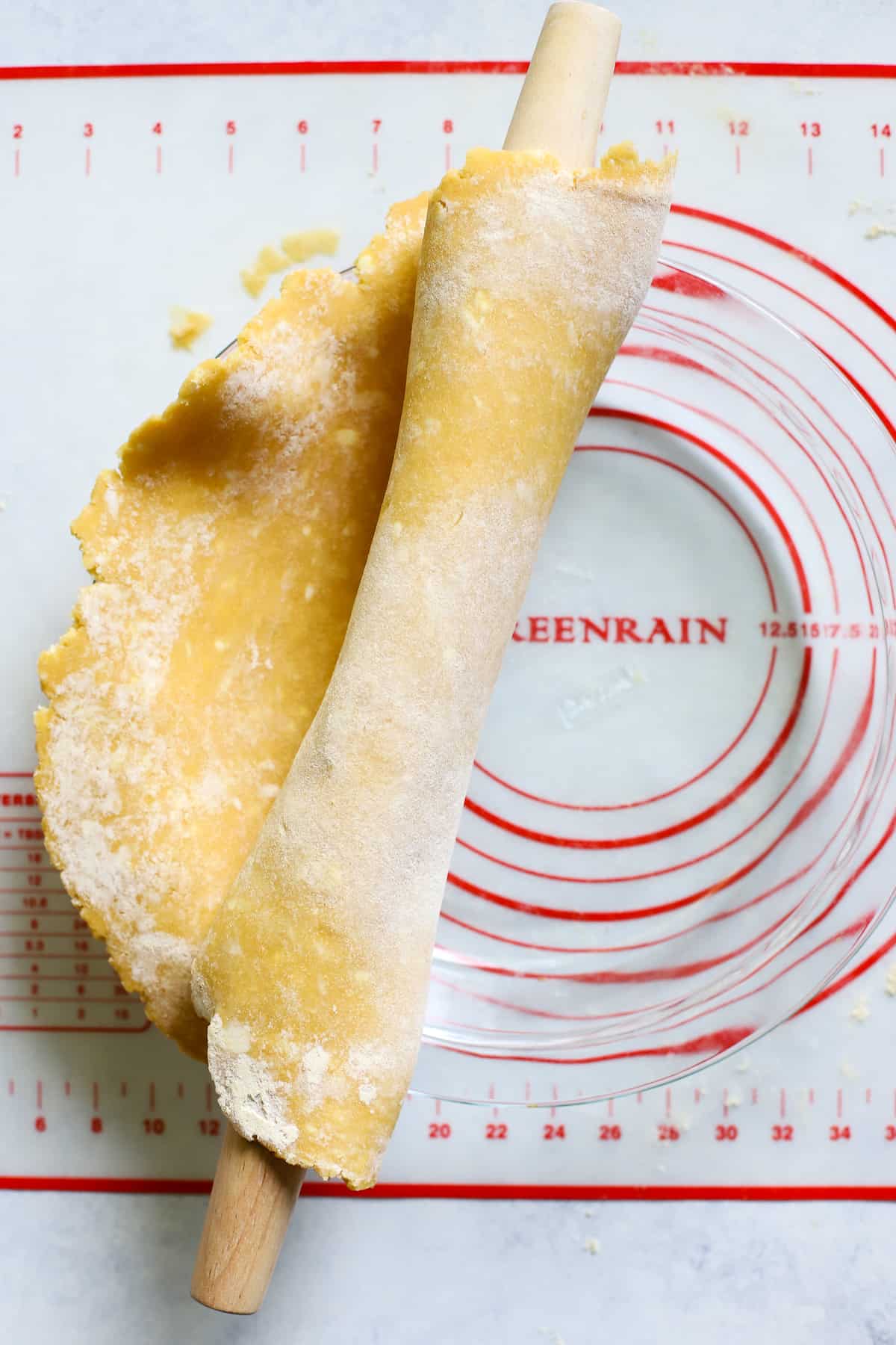 French rolling pin rolling chickpea flour pie crust into a clear glass pie dish, on pastry mat