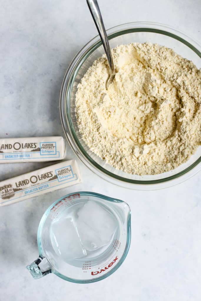 Chickpea flour in clear glass bowl, two sticks of butter, and ice water