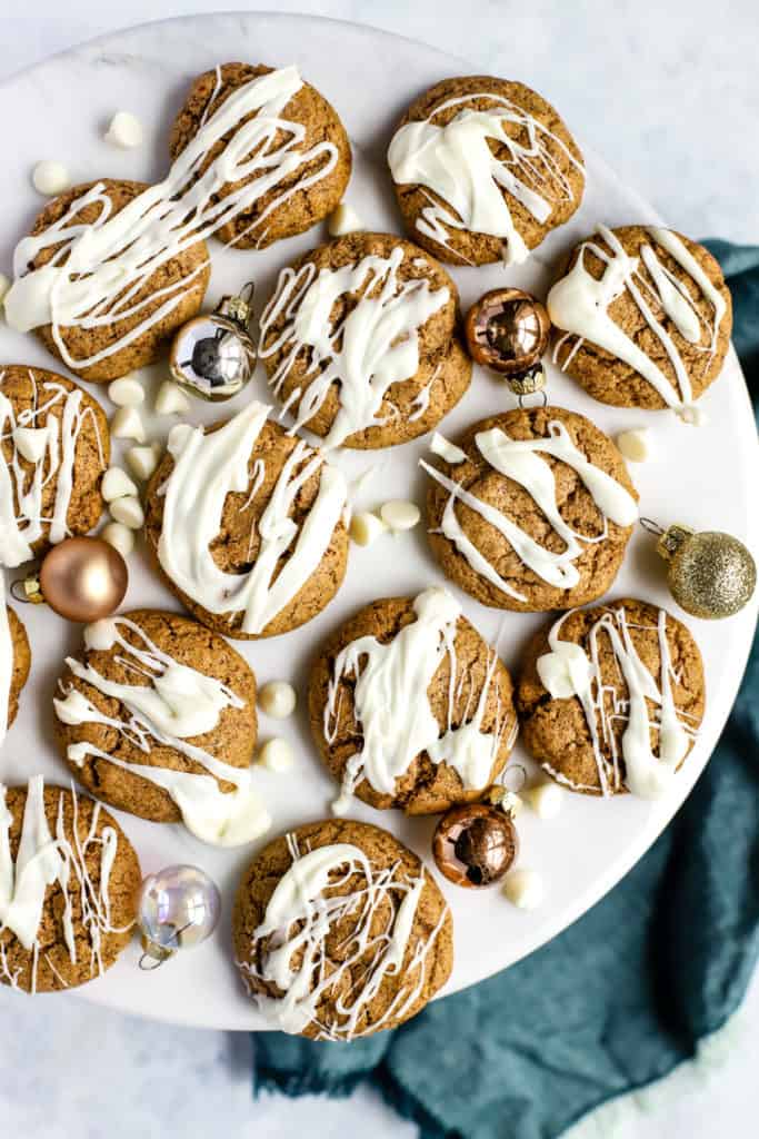 Flourless almond butter cookies, drizzled with white chocolate, on white marble surface with a few small ornaments and white chocolate chips sprinkled around