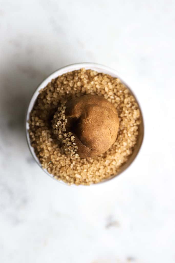 Ginger molasses cookie dough ball being rolled in coarse (turbinado) sugar in small white dish, on white marble surface