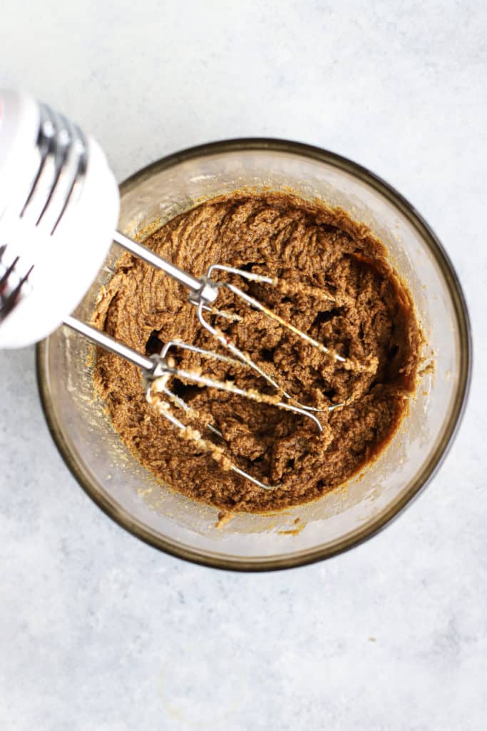 Wet ingredients for healthier ginger molasses cookies in glass bowl, with hand mixer beating the ingredients together