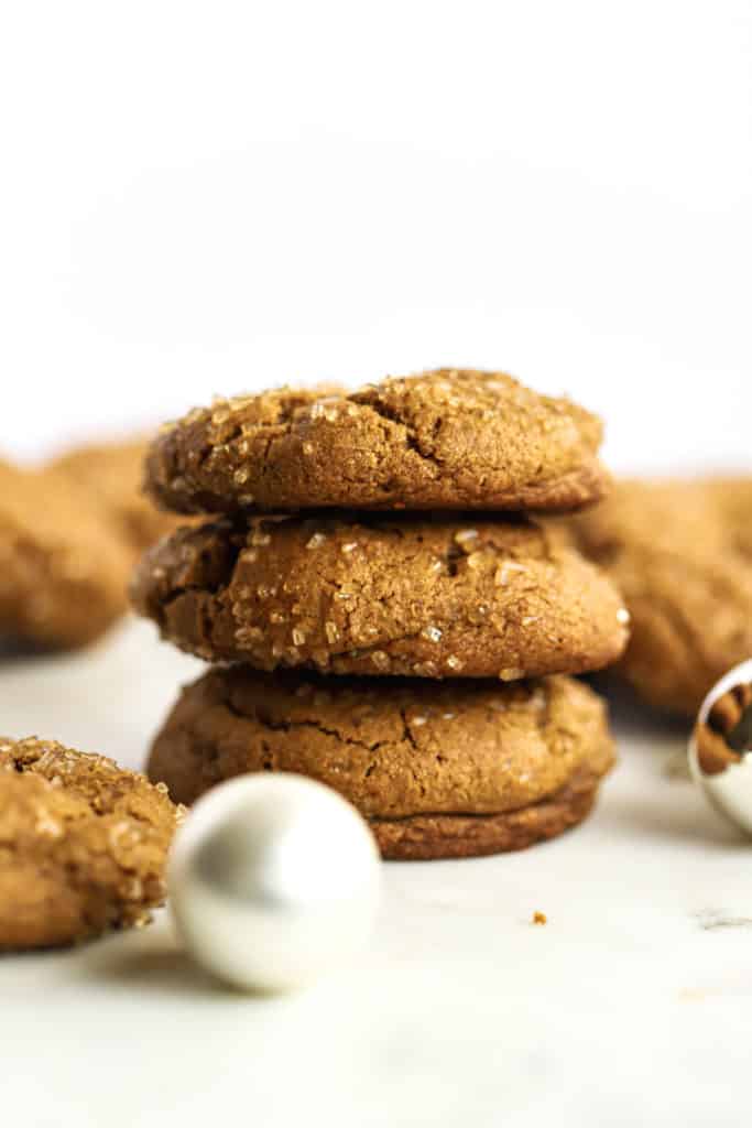 A stack of three healthier soft ginger molasses cookies, with more cookies in the background, on white marble surface with a few ornaments